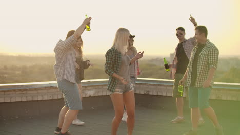 A-company-of-six-young-people-threw-a-party-with-beer.-Russian-girl-is-dancing-on-the-roof-with-her-friends-on-the-teamy-party.-She-dances-in-denim-shorts-and-green-plaid-shirts.